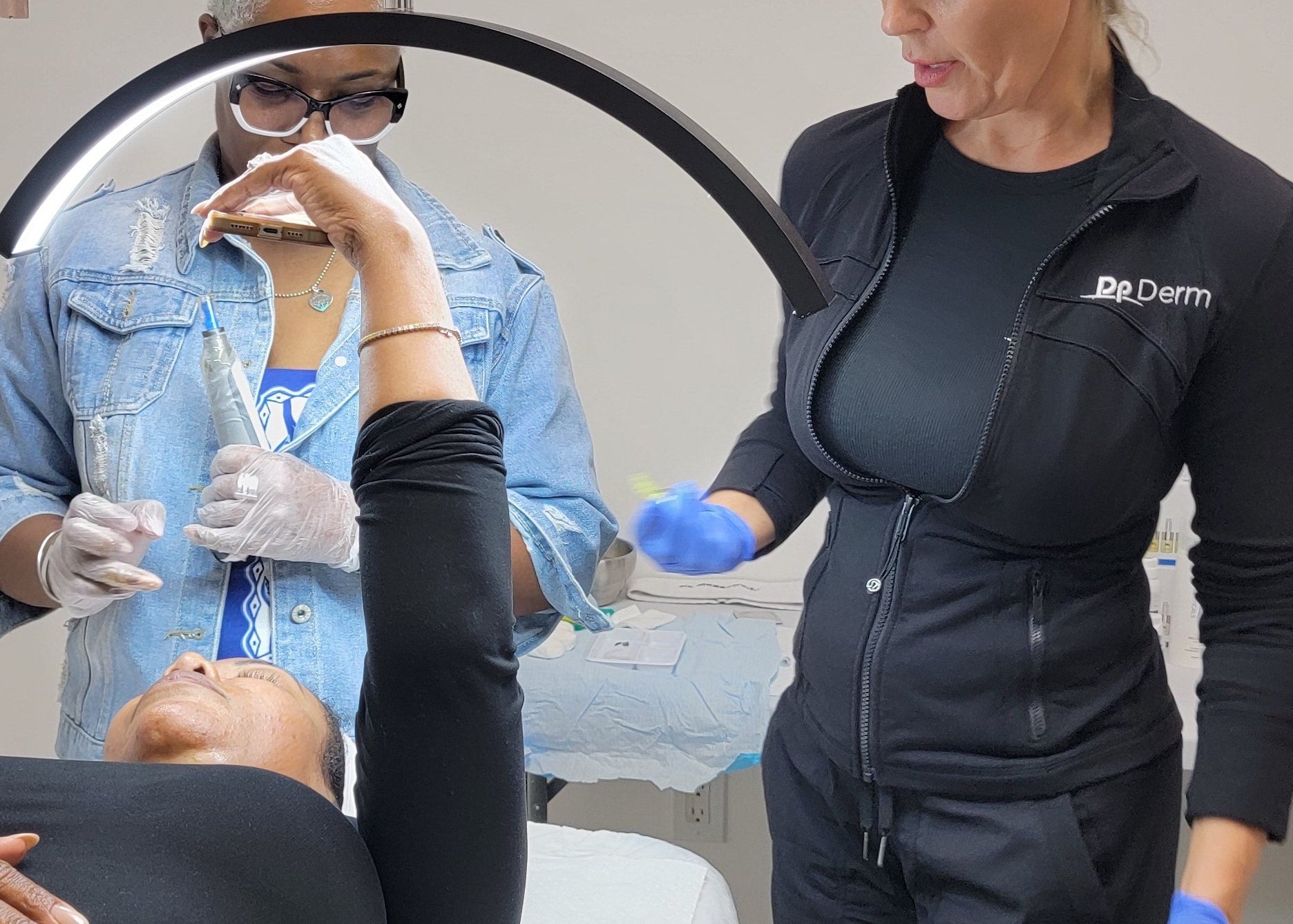 Two professionals in a medical or cosmetic setting, one wearing a black jumpsuit with 'Dr. Derm' branding, attending to a patient.